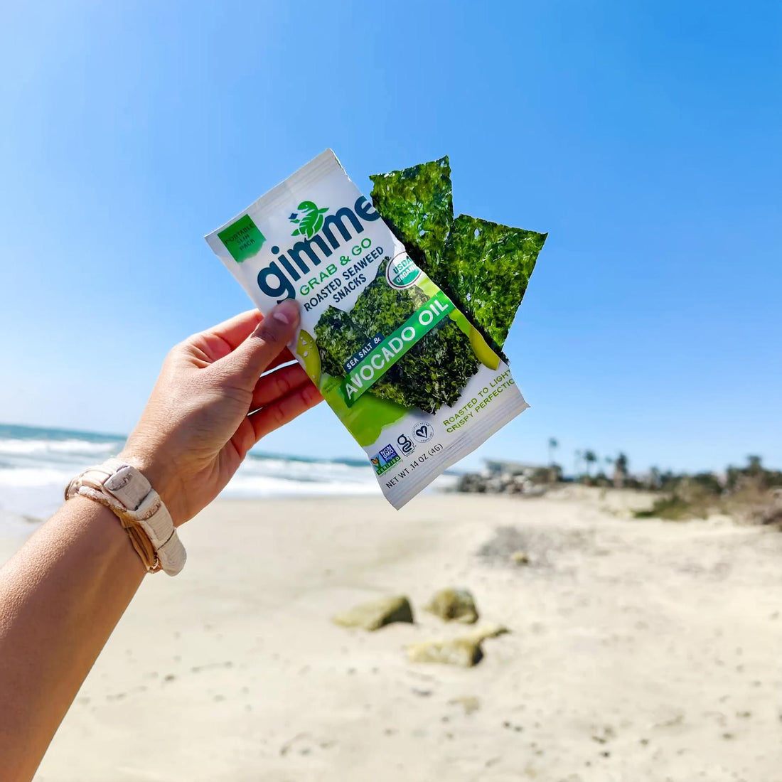 Image of a hand holding gimme avocado oil seaweed snack on a beach.
