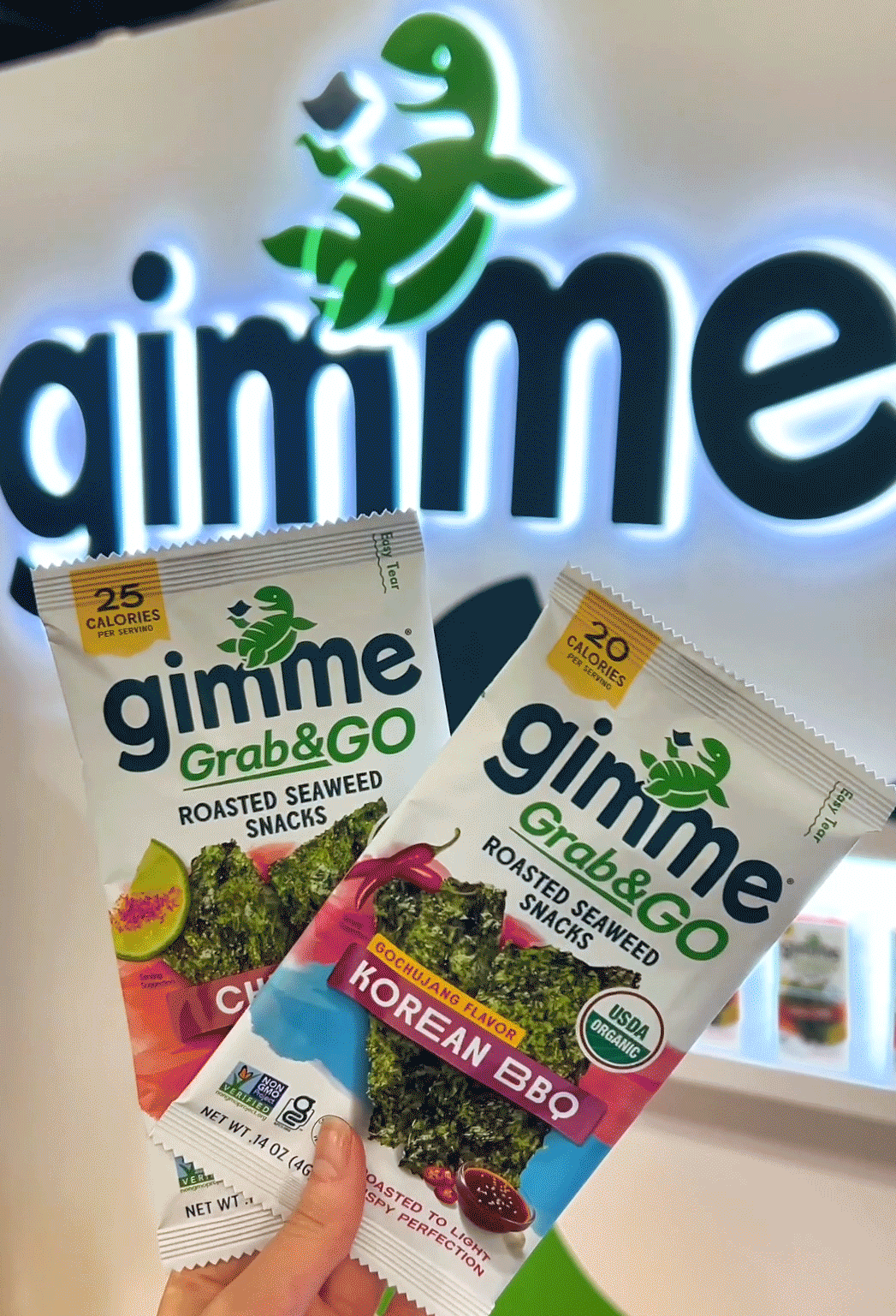 Single packs of Gimme Chili Lime Grab & Go Roasted Seaweed Snack and Gimme Korean BBQ Grab & Go Roasted Seaweed Snack held up in front of a Gimme Seaweed sign on a wall.