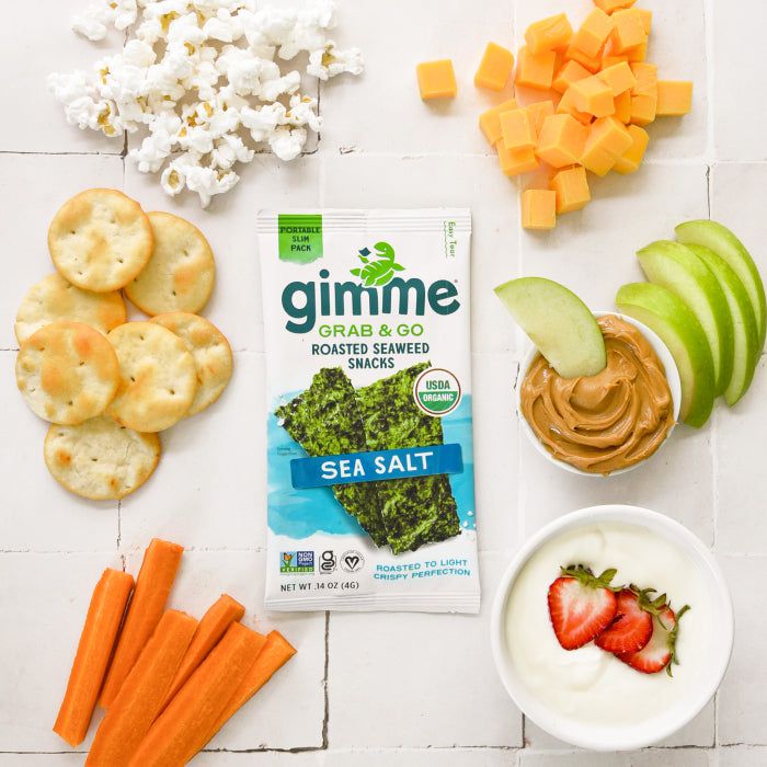 Top view of a variety of snacks, including popcorn, cheese cubes, apple slices with peanut butter, yogurt with strawberries, seaweed snacks, crackers, and carrot sticks arranged on a table.