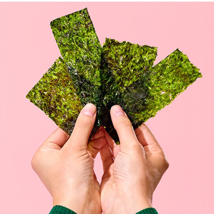 Hands holding several pieces of green seaweed snacks in front of a pink background.
