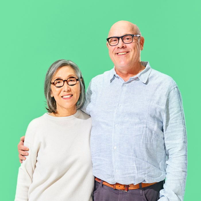 An older couple stands side by side smiling against a green background. The woman wears glasses and a light sweater, and the man wears glasses and a light blue shirt.