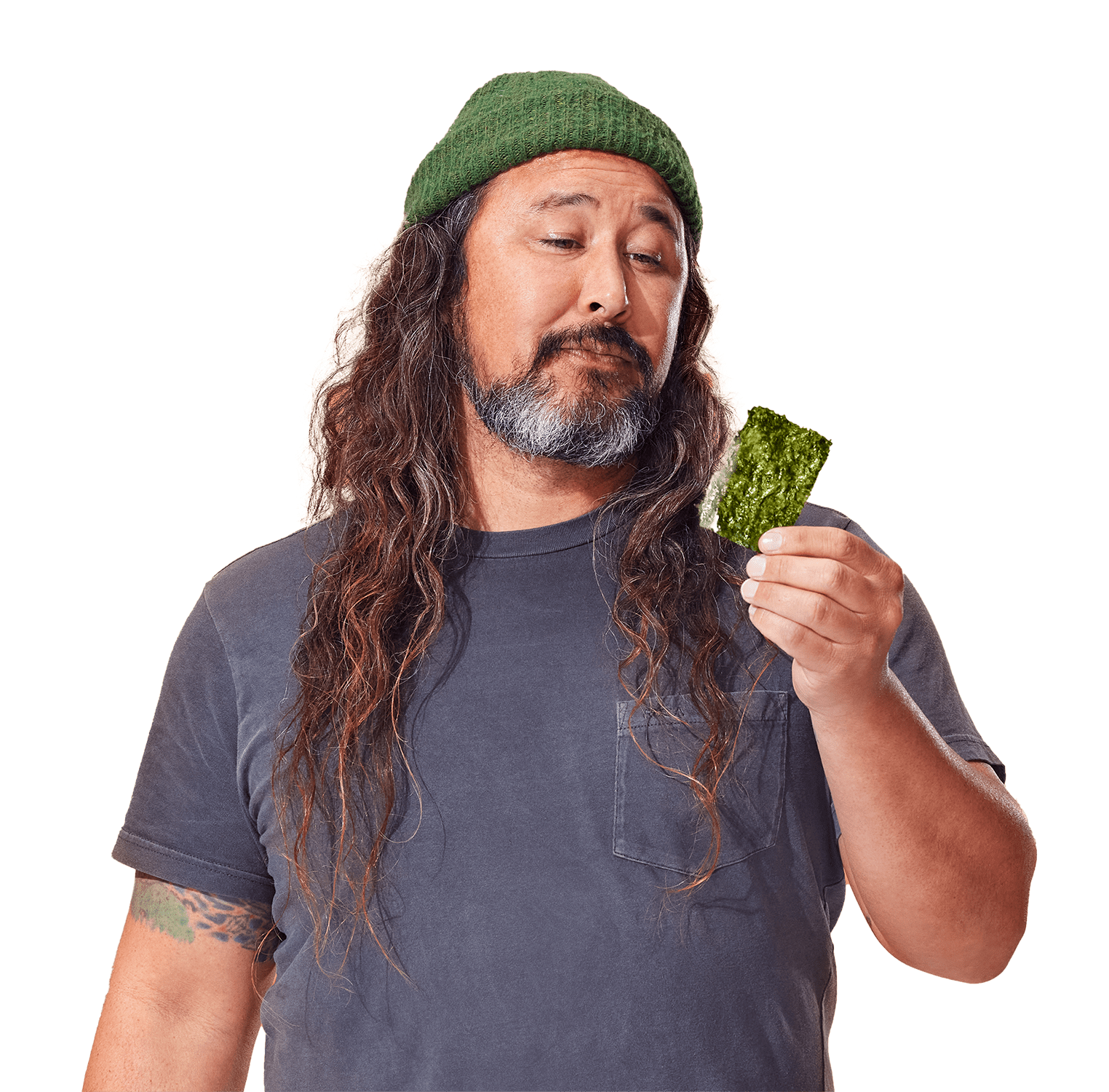 A man wearing a green beanie and gray shirt looks at a piece of green seaweed in his hand against an orange background.