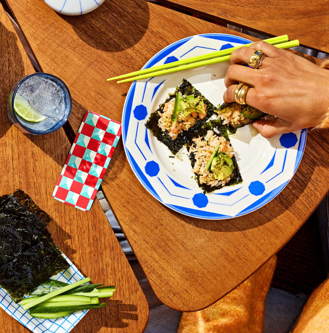 A hand with rings holding chopsticks, reaching for sushi on a blue and white plate. Seaweed, cucumber sticks, a drink with lime, and a red-green checkered packet are on a wooden table.