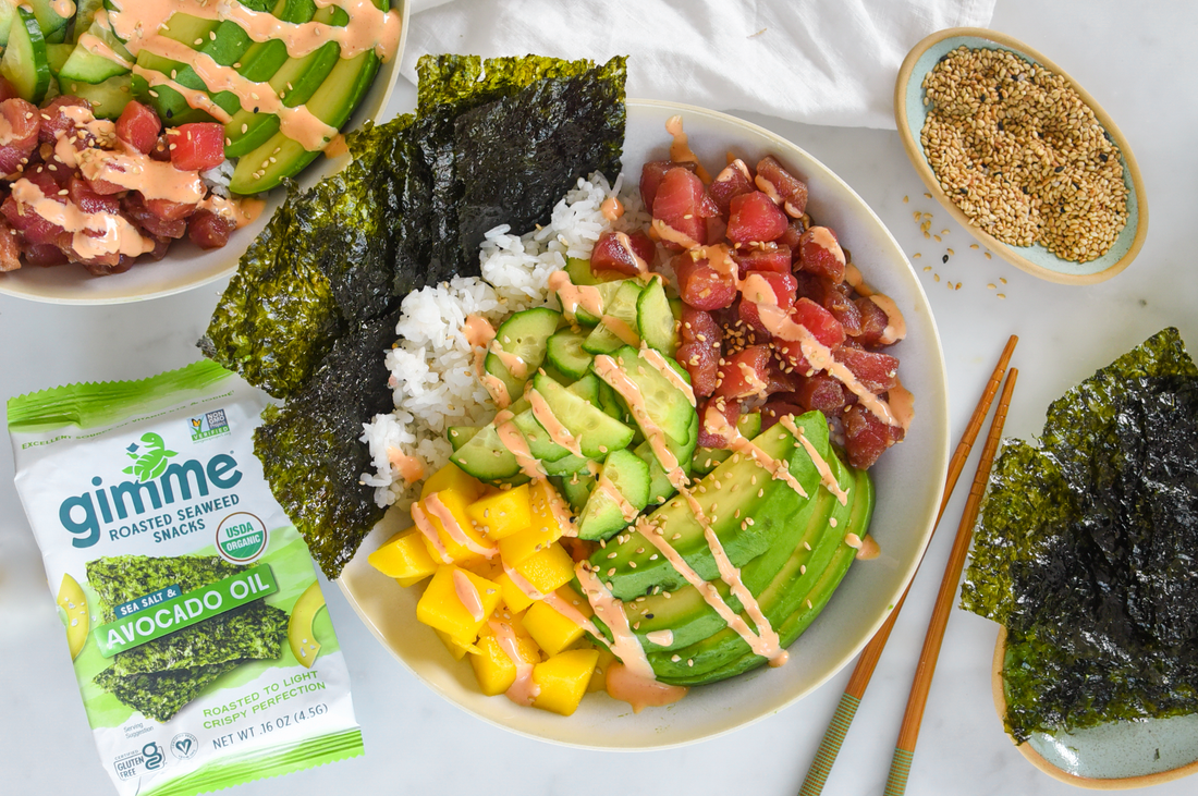 An image of a bowl of white rice topped with cubes of raw Ahi Tuna, avocado slices, mango pieces, cucumber, and drizzled sauce, accompanied by seaweed sheets, a packet of roasted seaweed, and a bowl of seeds.