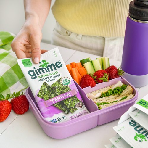 A lunch box with a sandwich, fruits and vegetables and gimme teriyaki seaweed snack.