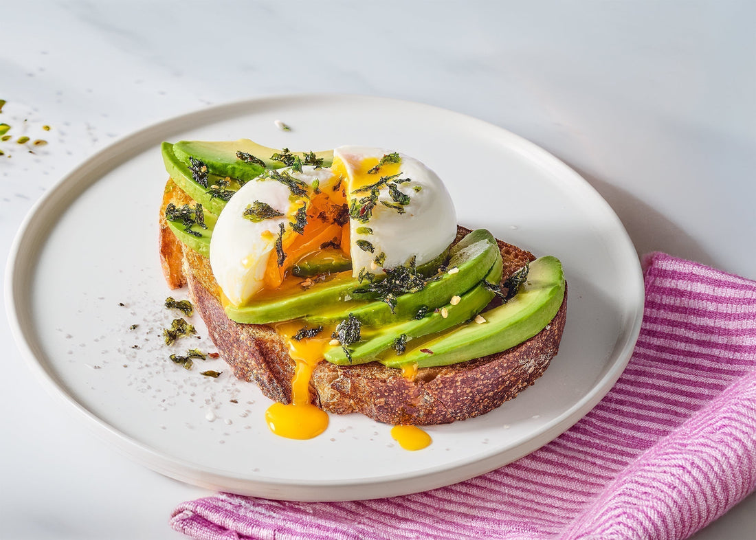 Avocado toast with poached eggs and Gimme K-Crisps Seaweed Topping on a white plate, placed on a pink and white striped towel.