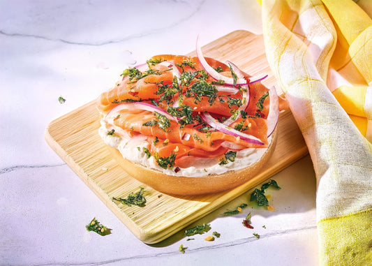 Smoked salmon and cream cheese on a bagel with Gimme K-Crisps Seaweed Topping sprinkled on top, sitting on a wooden board with a yellow cloth.