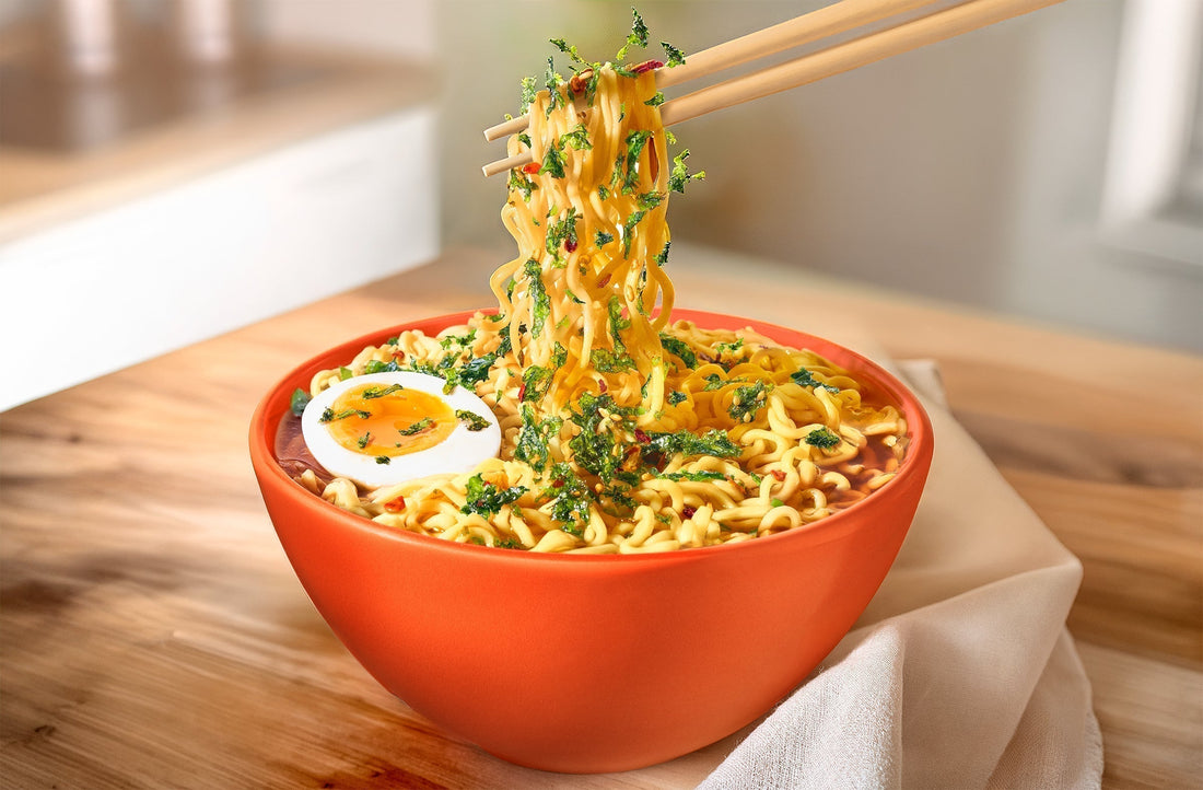Orange bowl of ramen noodles with an egg and Gimme K-Crisps Seaweed Topping, held by chopsticks on a wooden surface.