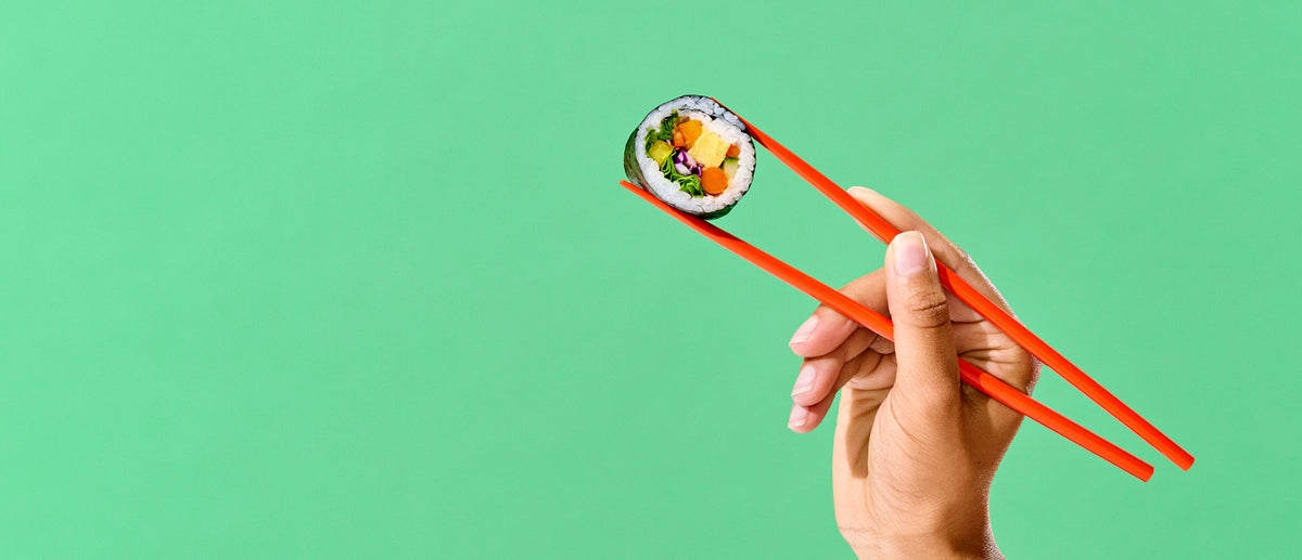 A hand holding red chopsticks grips a single piece of sushi with a green background.