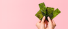 Hands holding several sheets of green seaweed snacks against a pink background.