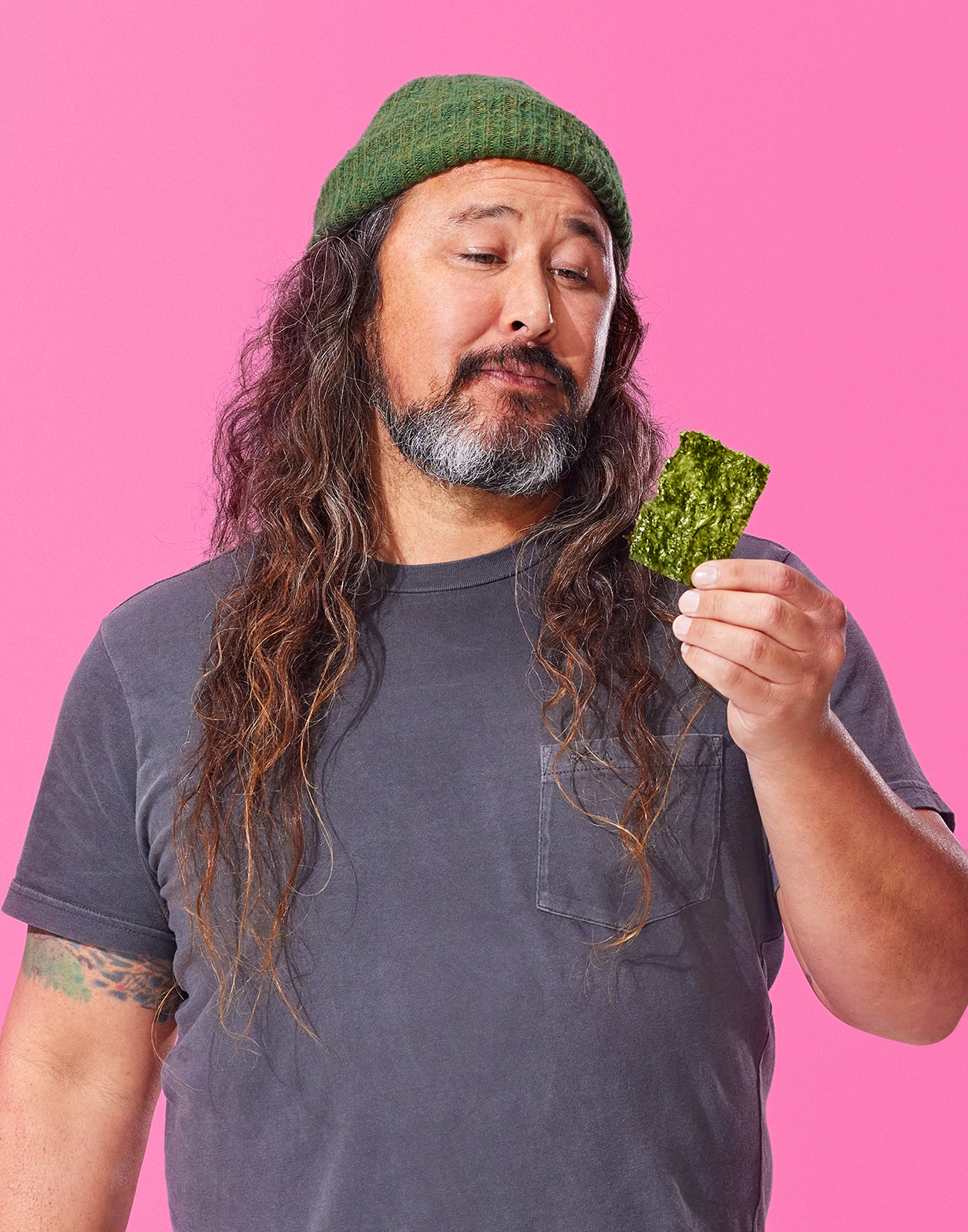Man with long hair and beard holding a piece of Gimme seaweed snack against a pink background