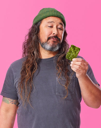 Man with long hair and beard holding a piece of Gimme seaweed snack against a pink background