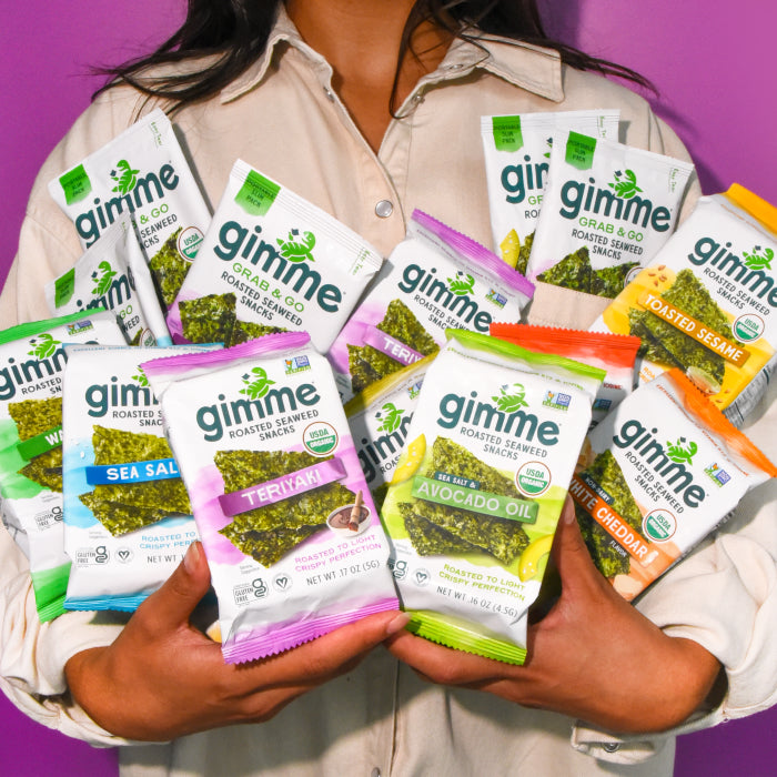 Person holding multiple packs of gimme roasted seaweed snacks with various flavors against a purple background.