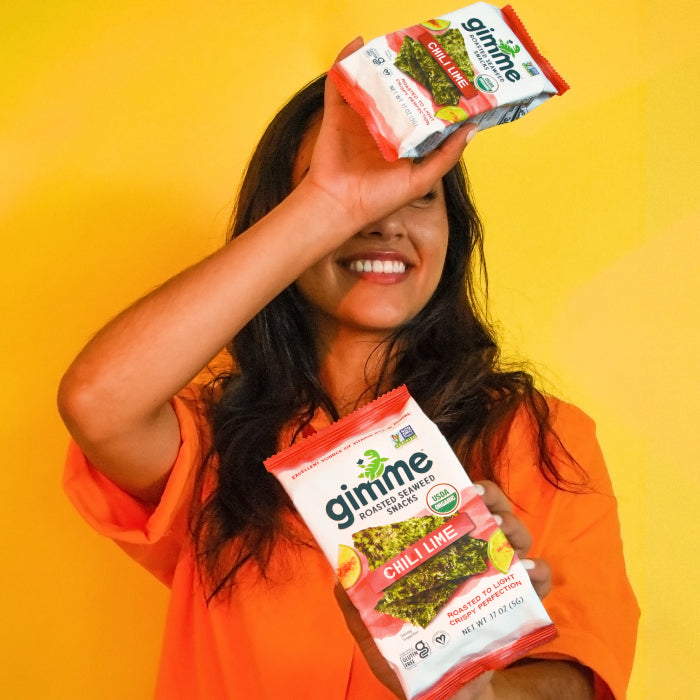 A person wearing an orange shirt holds two bags of gimme roasted seaweed snacks, one over their head and the other in front of their chest, against a yellow background.