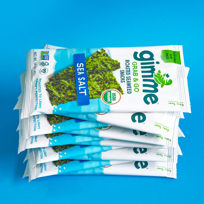 Stack of gimme Grab & Go Roasted Seaweed Snacks in Sea Salt flavor, displayed in a neat pile against a blue background. Packaging shows USDA Organic and Non-GMO Project Verified labels.