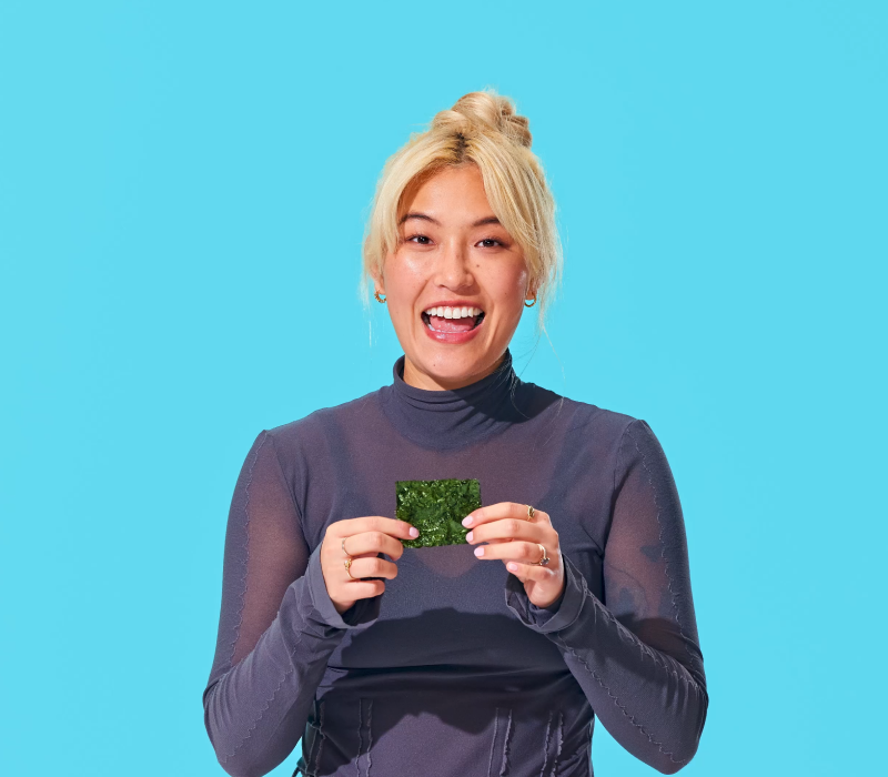 Person with blonde hair in a bun, wearing a long-sleeve dark shirt, holds and smiles at a piece of green seaweed against a solid turquoise background.