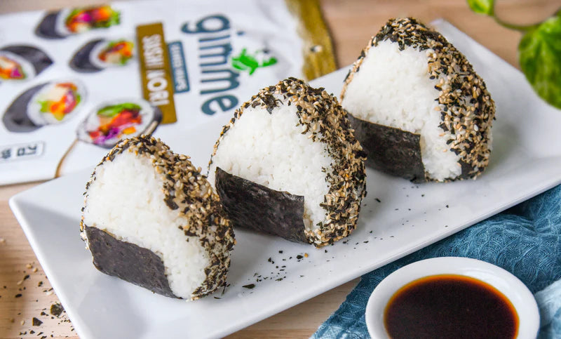 Three triangular rice balls with seaweed and sesame seed seasoning on a white plate, with a side dish of soy sauce.