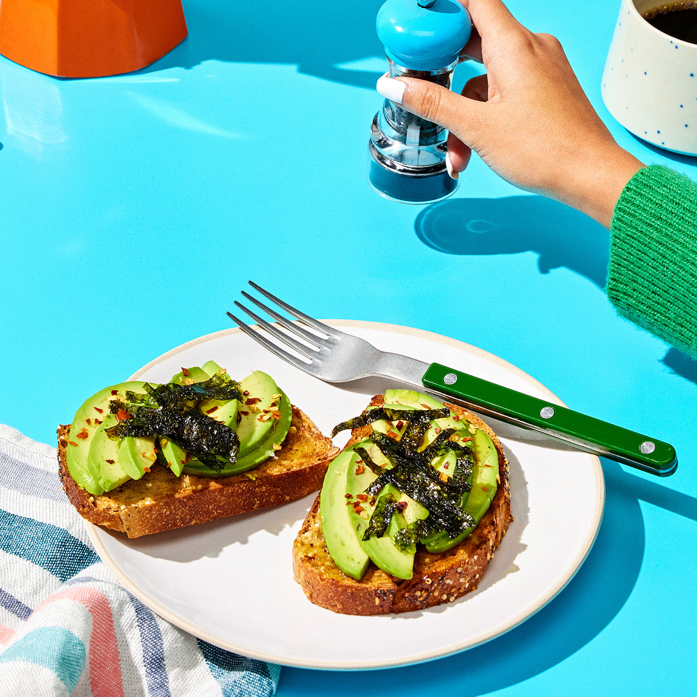 A person seasons avocado toast with a blue spice grinder while holding a fork on a plate with two pieces of toast on a light blue table. A coffee cup and cloth with stripes are nearby.