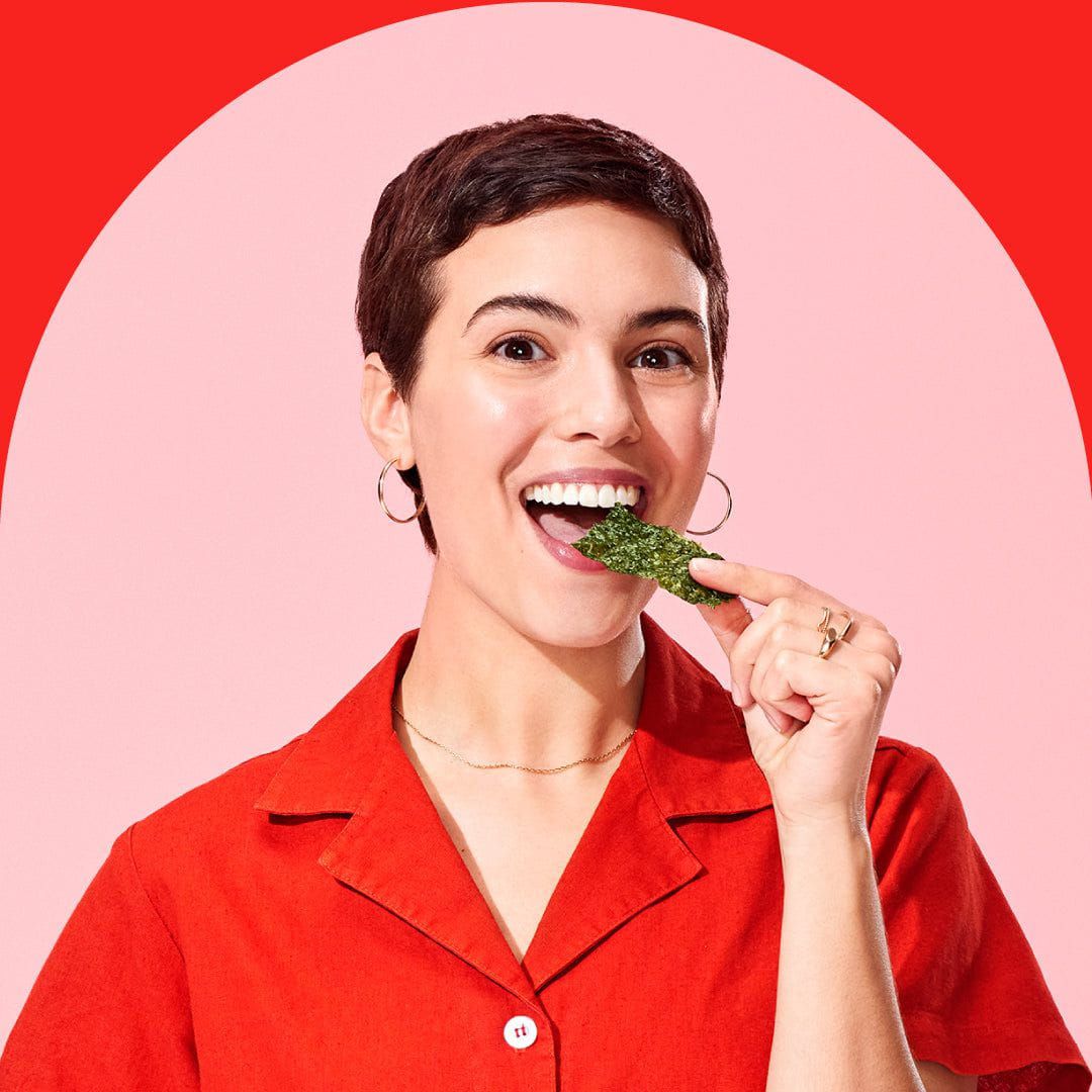 Person with short dark hair, wearing a red shirt, eating a green snack, and smiling against a red and pink background.