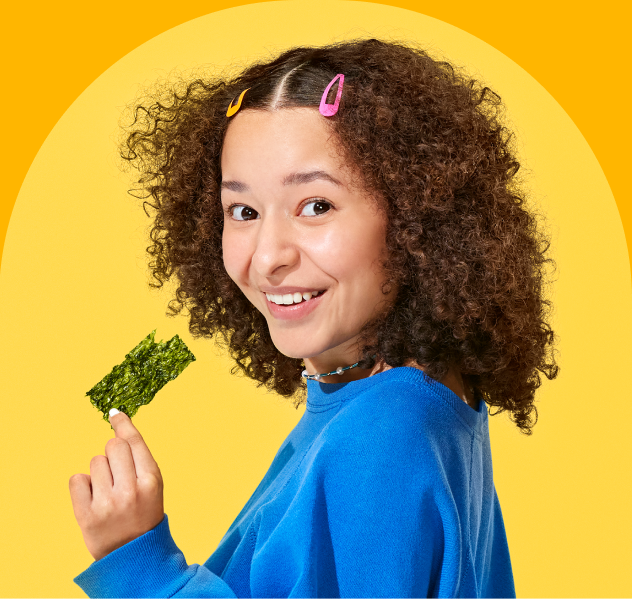 A person with curly hair and colorful hairpins holds a piece of seaweed while smiling, set against a bright yellow background.