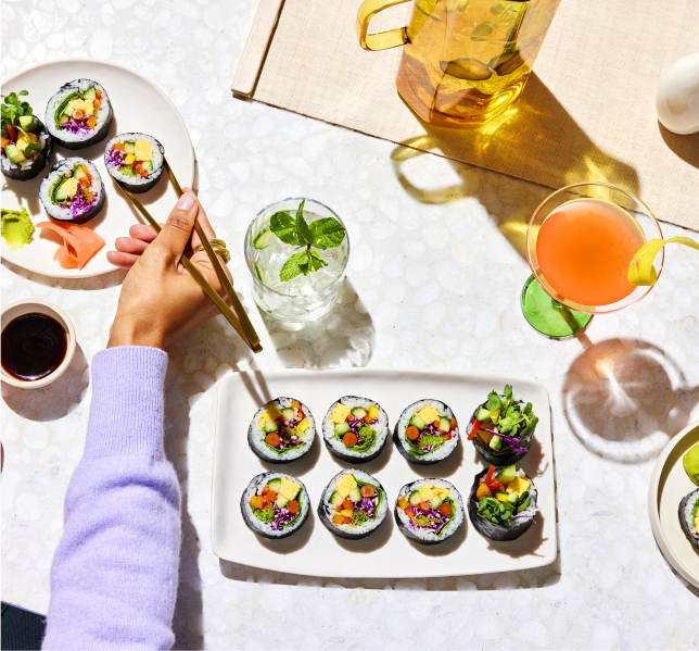 A person eating sushi with chopsticks over a white table set with various sushi rolls, a drink with mint leaves, a pink beverage in a cocktail glass, and a yellow pitcher.