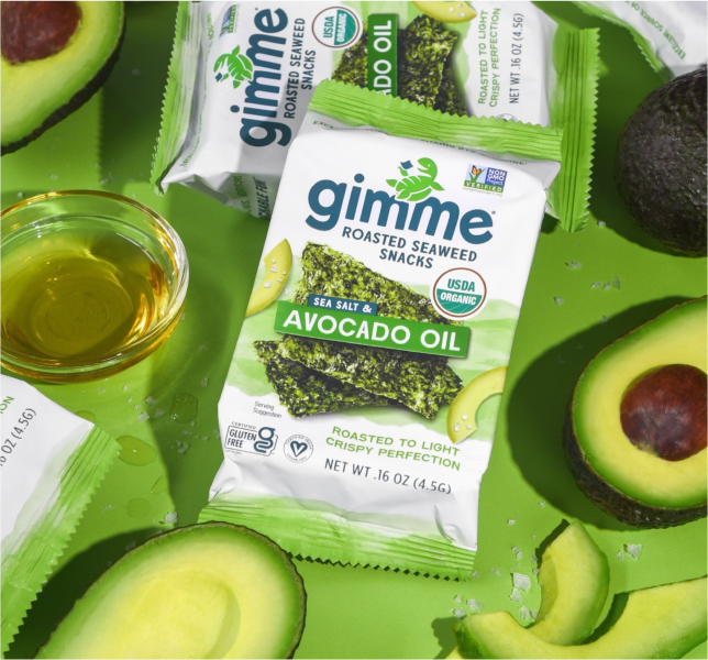 An arrangement of gimme brand roasted seaweed snacks in green packaging, with sliced avocados and a small bowl of avocado oil on a bright green surface.