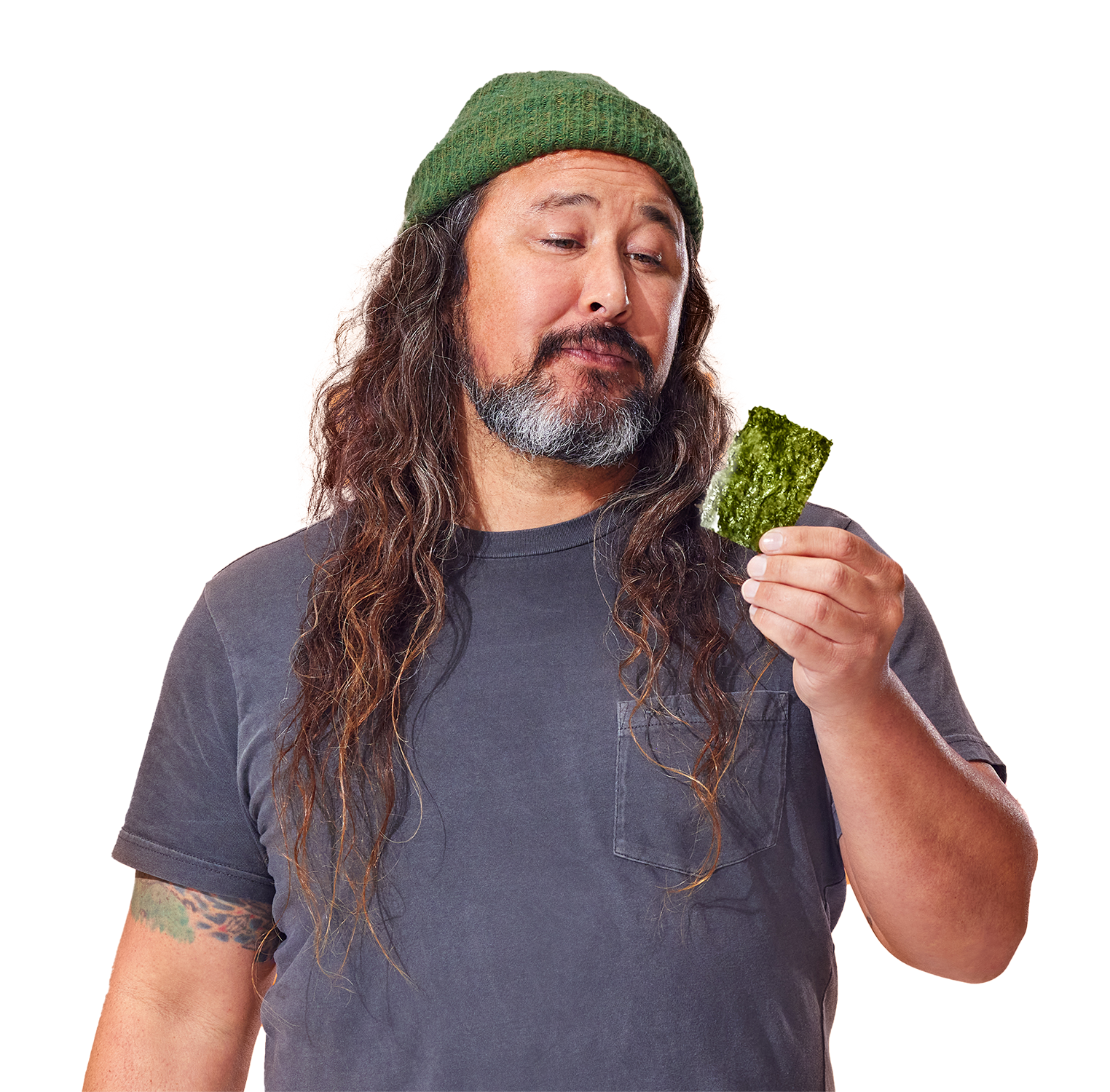 A man wearing a green beanie and gray shirt looks at a piece of green seaweed in his hand against an orange background.