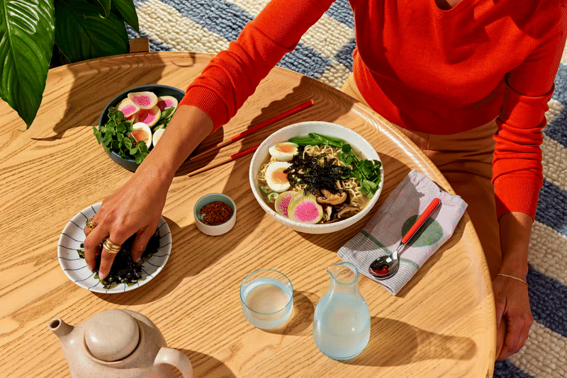 Person in a red shirt reaches for seaweed from a plate next to a bowl of ramen, with garnishes, a teapot, and glasses of water on a wooden table.