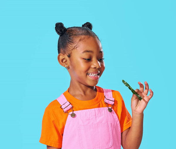 A young child with two buns in their hair, wearing a bright orange shirt and pink overalls, smiles while holding a small lizard in their hand against a light blue background.