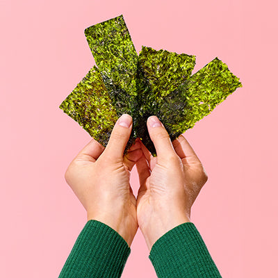 Hands holding several sheets of green seaweed snacks against a pink background.