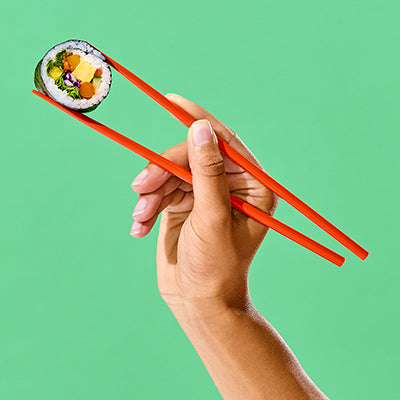 A hand holding red chopsticks grips a single piece of sushi with a green background.