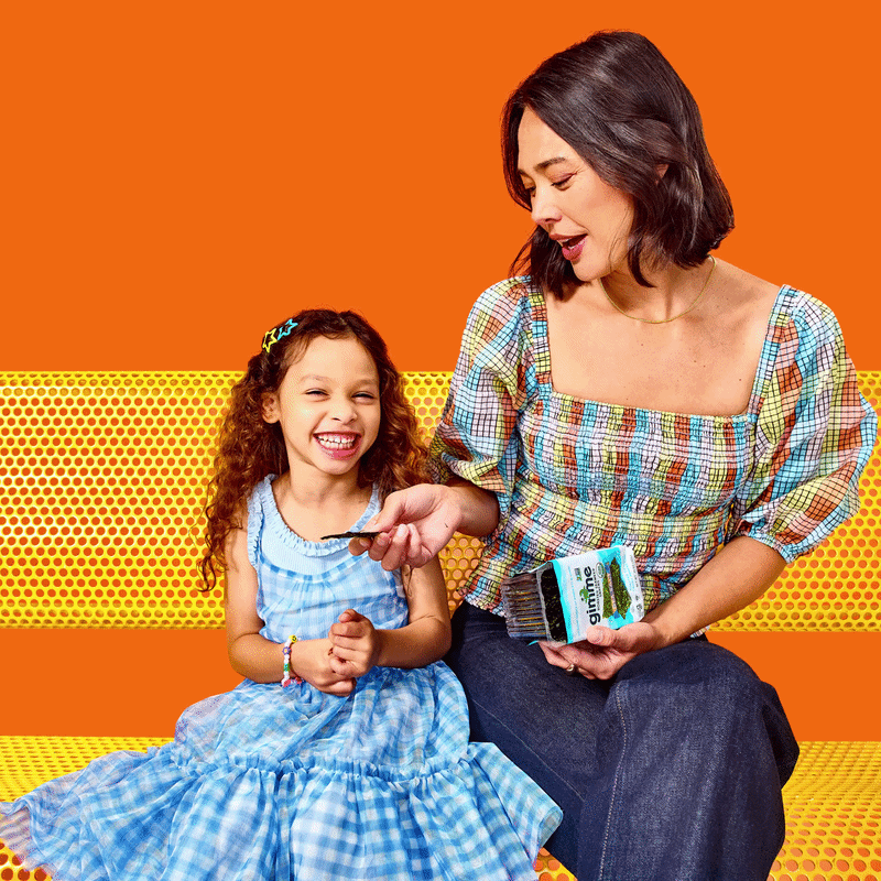 Mom handing her child a Gimme seaweed snack while sitting on a yellow bench 