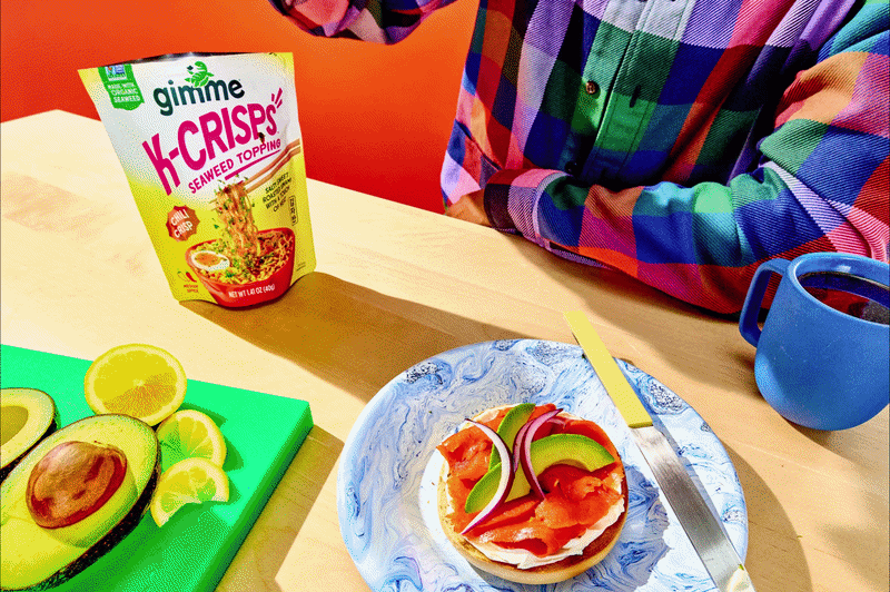 A person wearing a plaid shirt sprinkling gimme K-Crisp seaweed topping on top of a bagel with avocado slices.