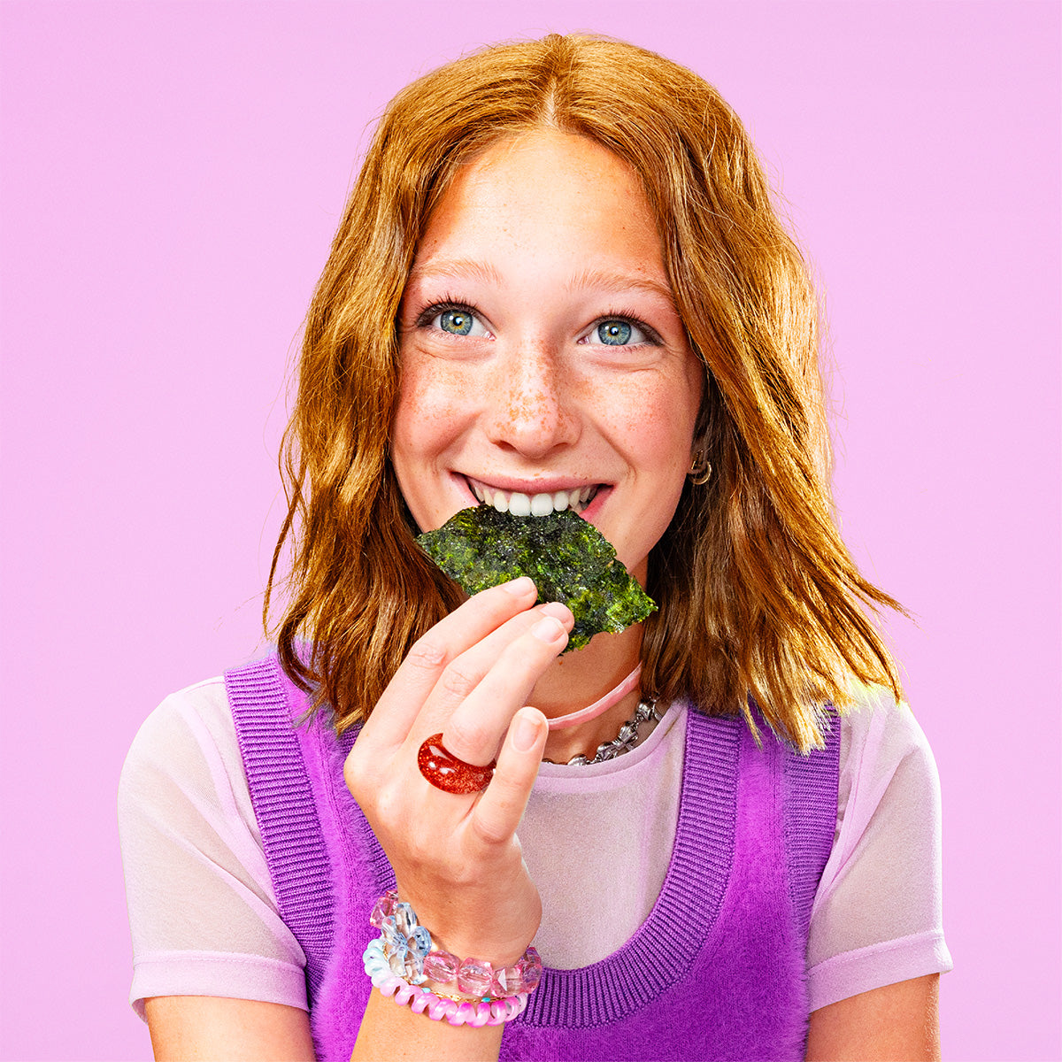 Woman eating a piece of seaweed snack against a pink background