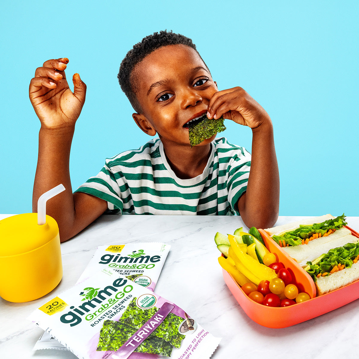 Child eating a Gimme seaweed snack with a colorful tray of vegetables on a blue background