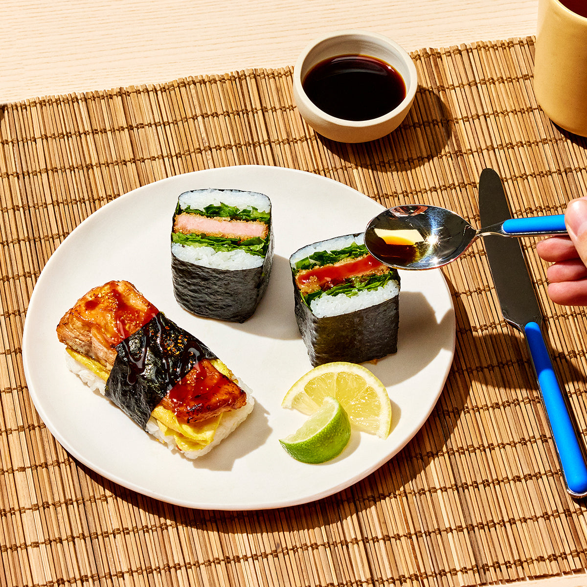 A plate with three pieces of sushi, including a musubi, accompanied by lime wedges. A hand drizzles sauce from a spoon, with chopsticks, a knife, and a cup of soy sauce nearby.